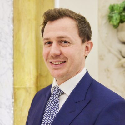 A professional photograph of Oliver Stokes in a suit and tie smiling.